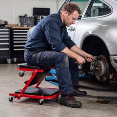 Man sitting on a red rolling creeper seat garage chair working on a car wheel