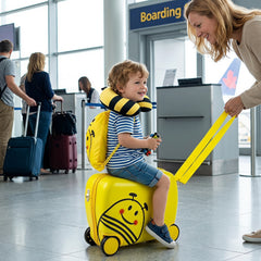 A child sits on the yellow kids luggage set with a bee design, with a neck pillow