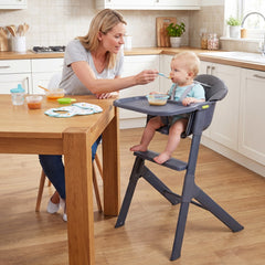 The high chair in a kitchen with a baby sitting in it while being fed by an adult at a table