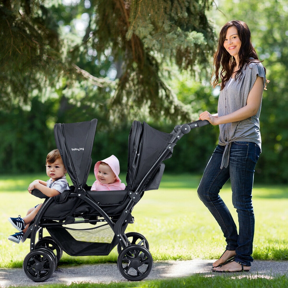 A woman pushes a black double pushchair with two babies sitting in it, outdoors in a park