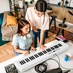 Two women playing the electronic keyboard in a bright room with musical instruments in the background