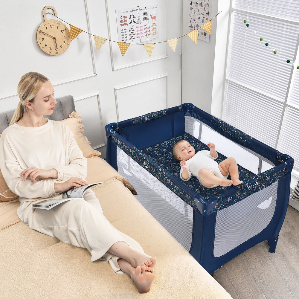 Mum sitting on a bed next to the blue baby playpen with a baby inside