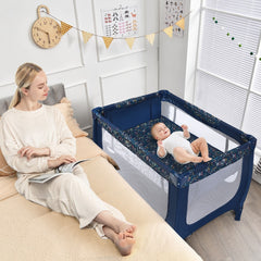 Mum sitting on a bed next to the blue baby playpen with a baby inside