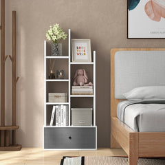 A white wooden bookcase with a grey drawer is shown in a bedroom setting next to a bed