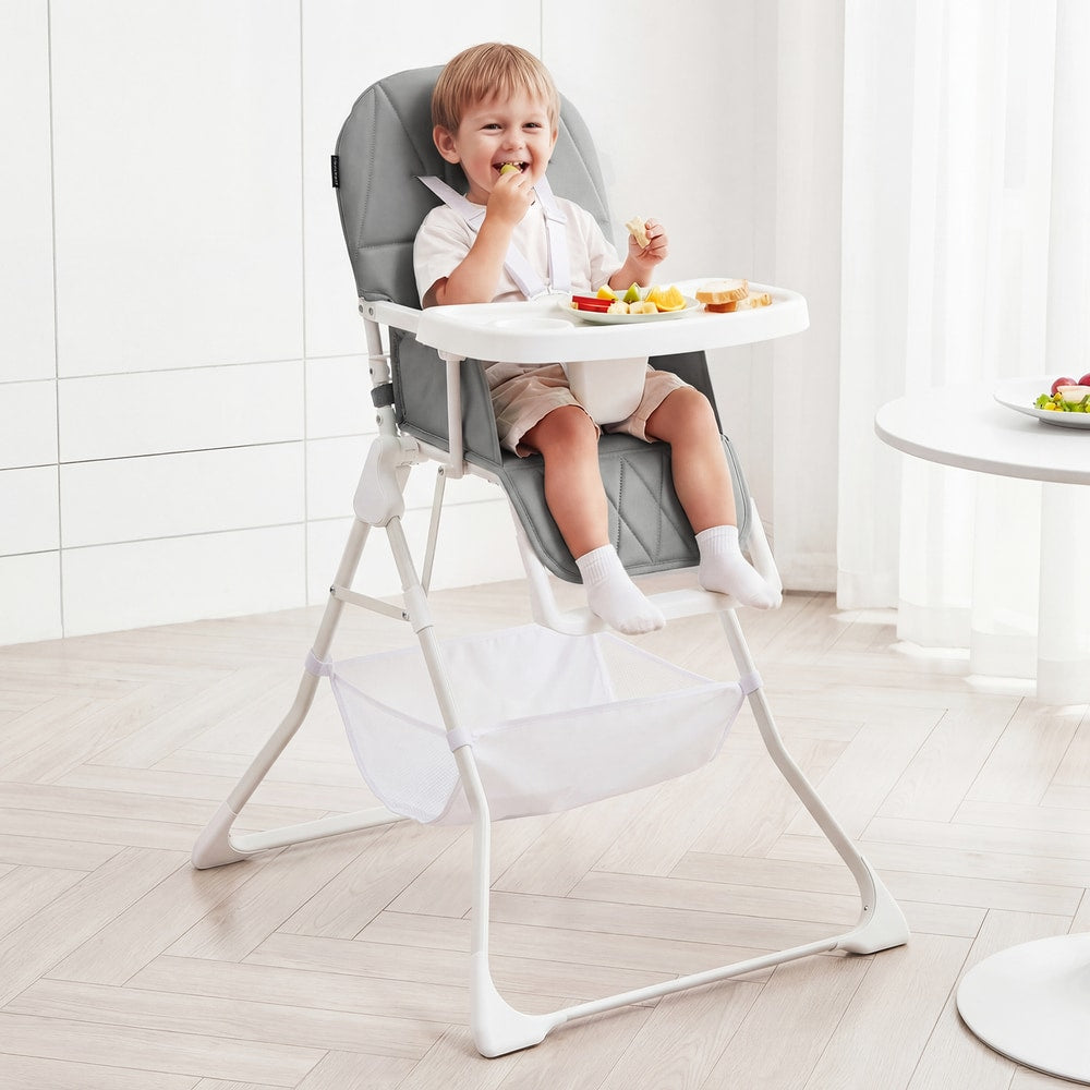 A toddler eats food while sitting in the grey portable high chair at a dining table
