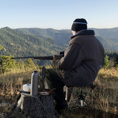 The camping stool in use by a hunter sitting on a tree stump in a mountainous landscape