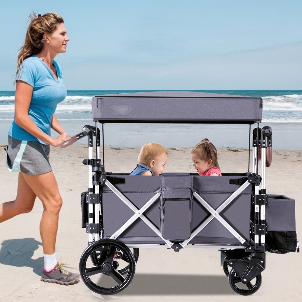A woman jogs with a grey double stroller wagon containing two babies on a sandy beach