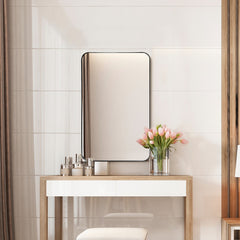 The bathroom mirror with rounded corners is shown above a vanity table with toiletries and flowers