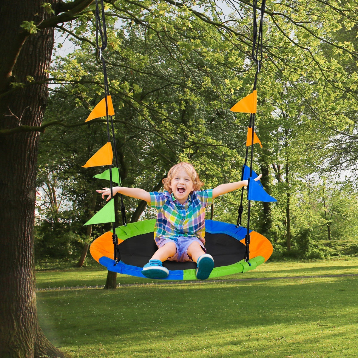 A happy child sitting on the round saucer swing set hanging from a tree in a green park