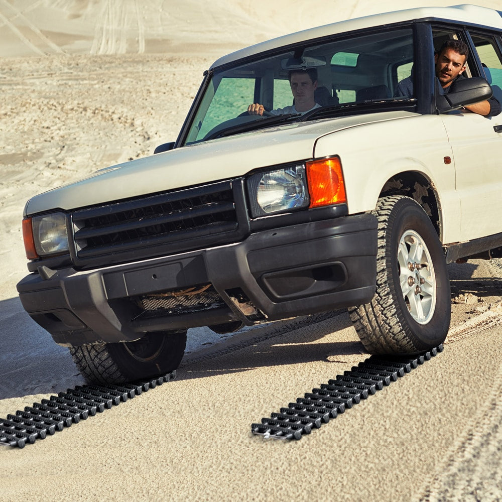 The recovery tracks placed under the front wheels of a white off-road vehicle on sandy terrain