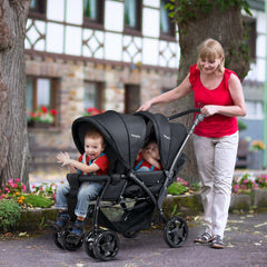 A woman pushes a black double pushchair with two babies sitting in it, on a paved path near buildings
