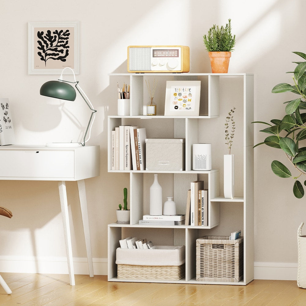 A white geometric bookcase filled with books and decor, placed next to a desk and sofa