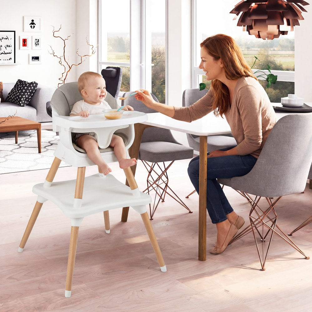 A baby is fed by a woman while sitting in the grey convertible high chair at a dining table