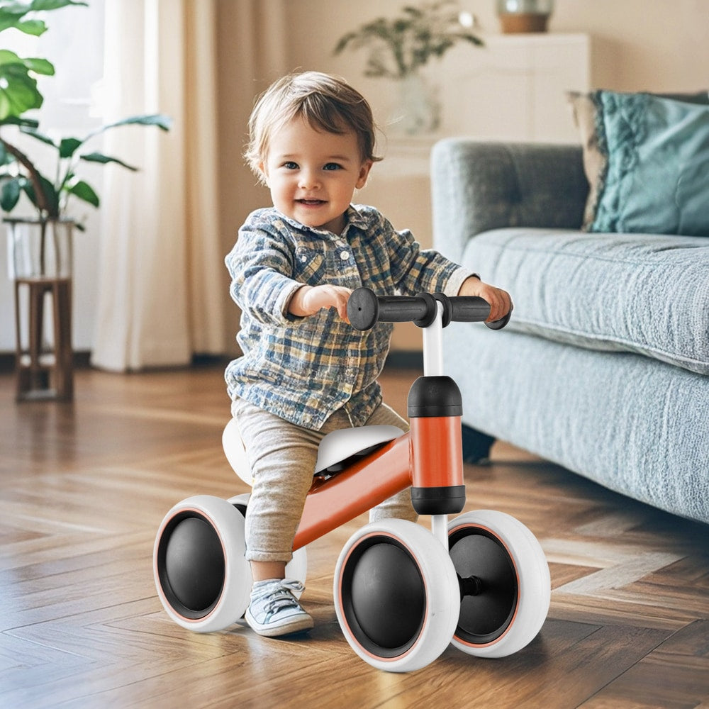A smiling toddler sits on the orange balance bike indoors in a living room