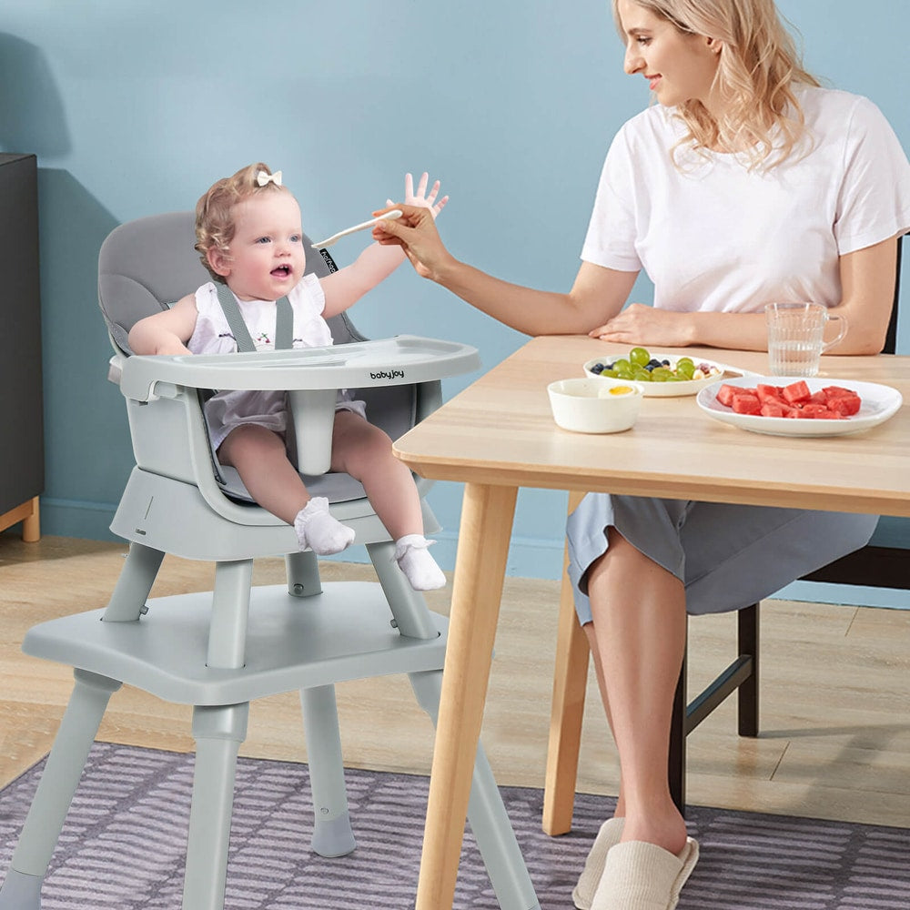 A baby sits in the grey high chair while a woman feeds her at a table
