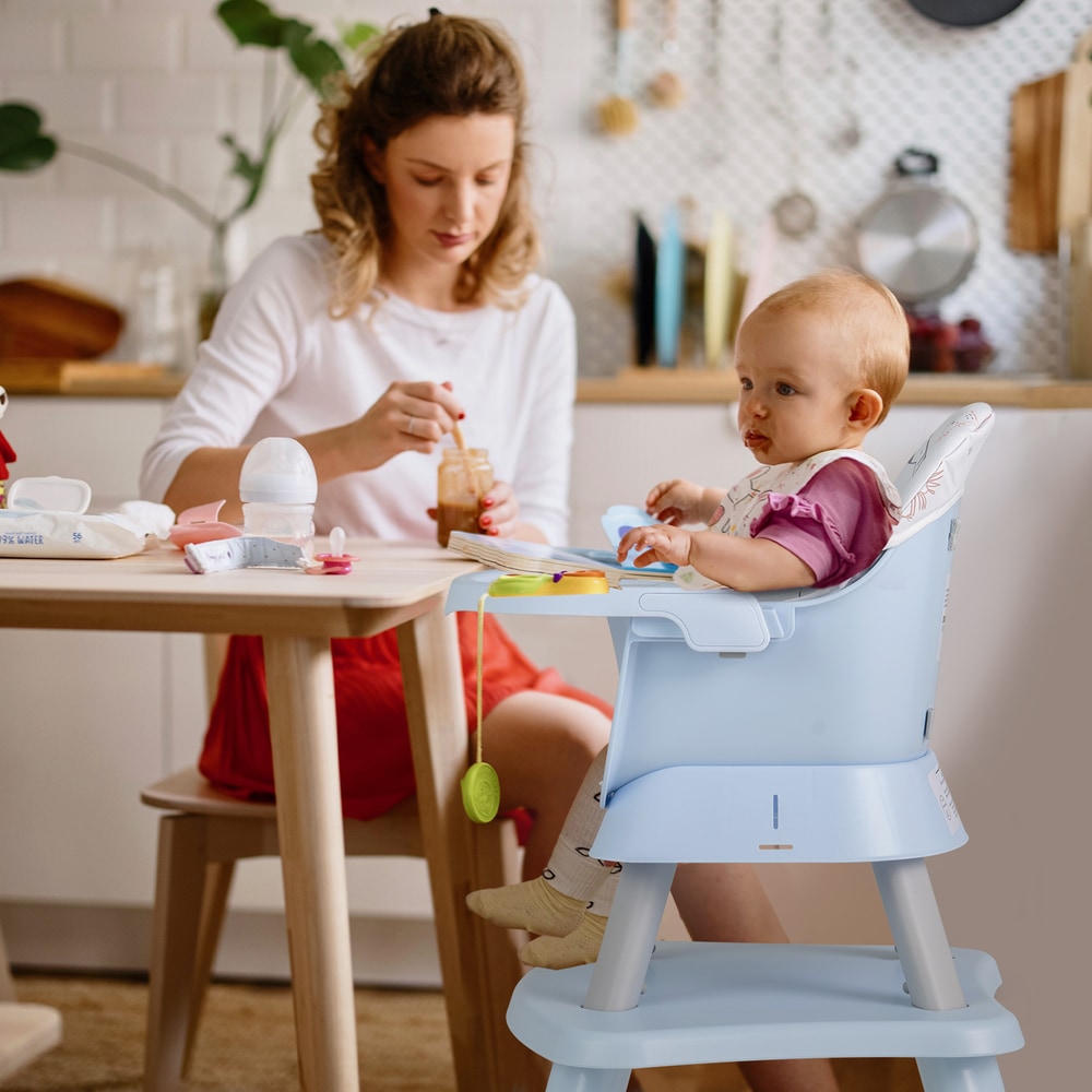 A woman feeding a baby in the blue high chair, which is set up as a booster seat at a table