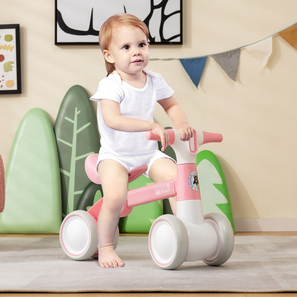 A toddler sits on the pink balance bike in a room with cartoon trees