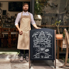 A man in an apron stands next to a double sided A frame chalkboard sign outside a shop