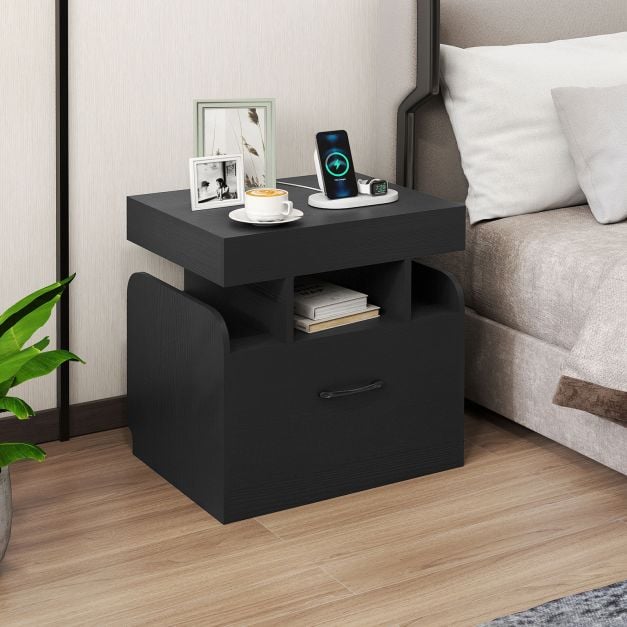 Black bedside table with a drawer, open shelf, and charging station next to a bed