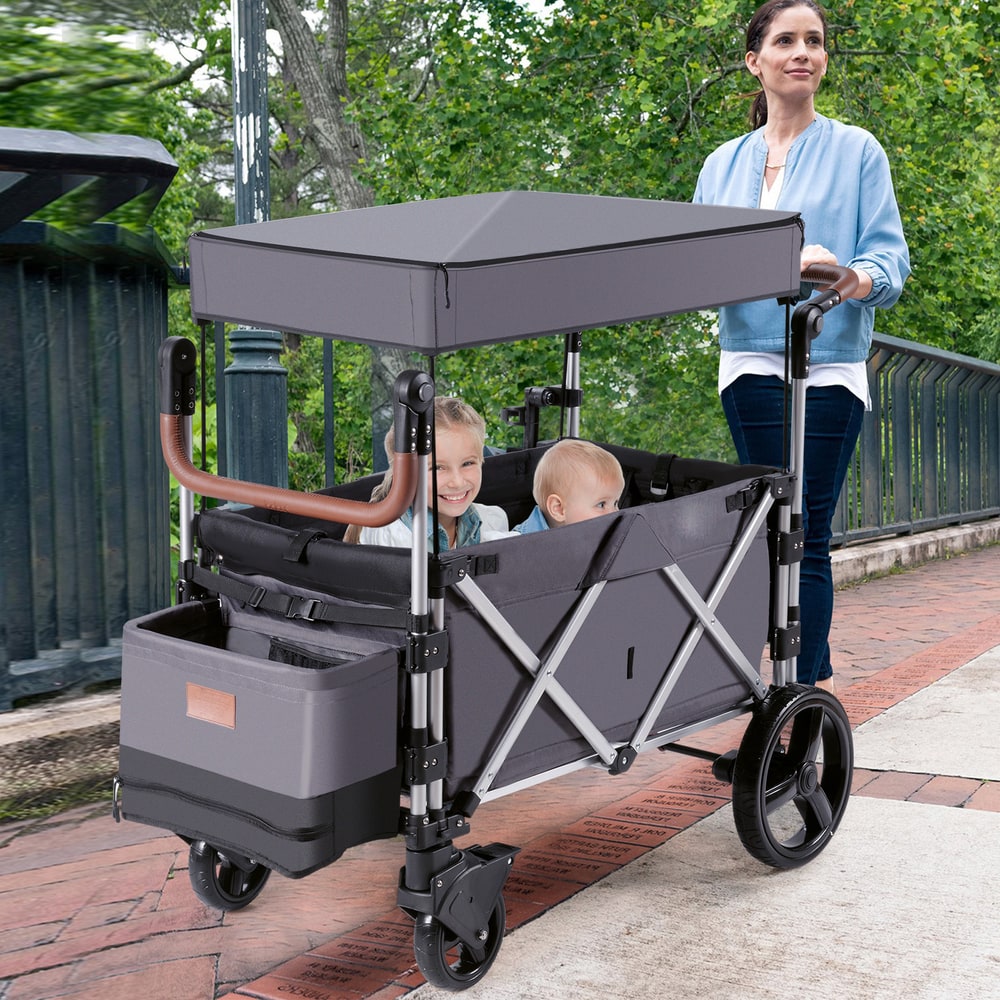 A woman pushes a grey double stroller wagon with two young children inside along a paved path