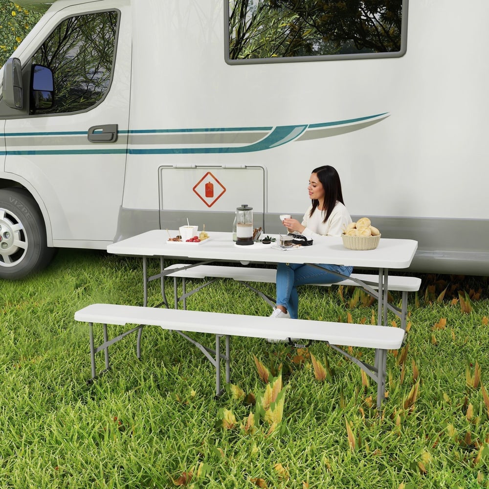 A woman sits at the white folding picnic table and bench set next to a campervan in a grassy area