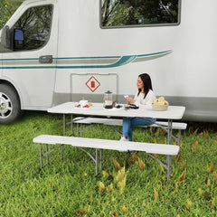 A woman sits at the white folding picnic table and bench set next to a campervan in a grassy area
