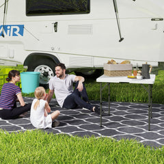The folding camping table set up on grass next to a caravan with people sitting around it